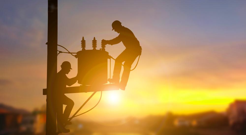 Two electrical engineers are seen in silhouette as they work on a power transformer.