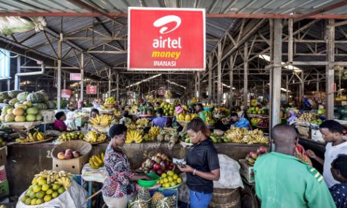 An advertisement for a mobile phone money transfer company hangs over stalls of fruit and vegetables at a covered market in Kigali, Rwanda. Photo: Panos/Sven Torfinn