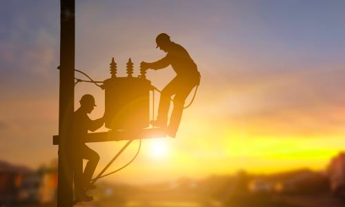 Two electrical engineers are seen in silhouette as they work on a power transformer.